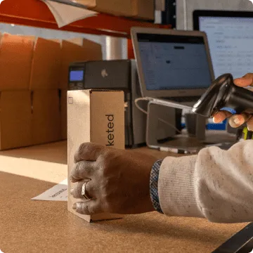 Eine linke Hand stellt eine Kartonschachtel mit dem komplett erneuerten Smartphone auf einen Tisch
